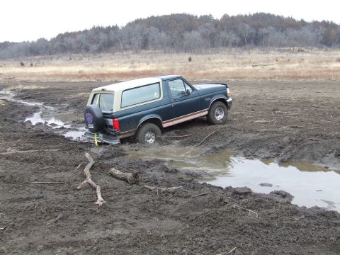 1994 Ford Bronco - A dark green Ford Bronco with a tan top is stuck in deep mud with trees in the background.