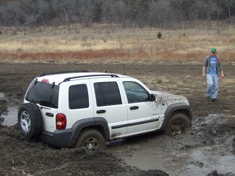 1994 Ford Bronco - A white Jeep Liberty is stuck in deep mud, with a person standing nearby in a rural, muddy landscape.