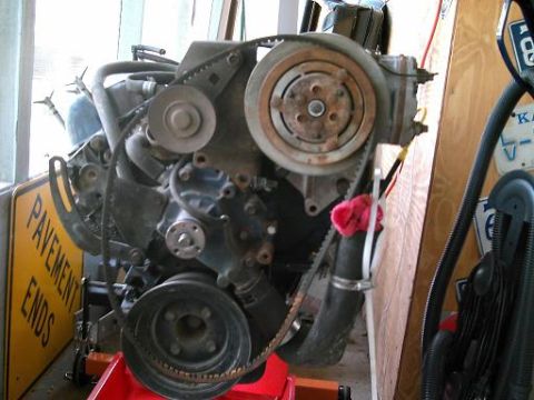 1986 Ford Bronco - Engine assembly with belts and pulleys, mounted on a red stand, with a 'PAVEMENT ENDS' sign in the background.