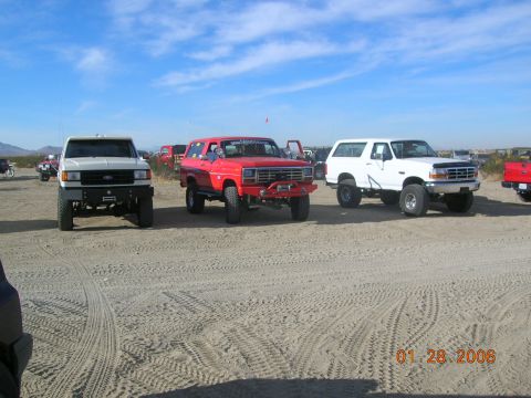 1985 Ford Bronco - Three SUVs parked on a sandy area under a blue sky with scattered clouds.