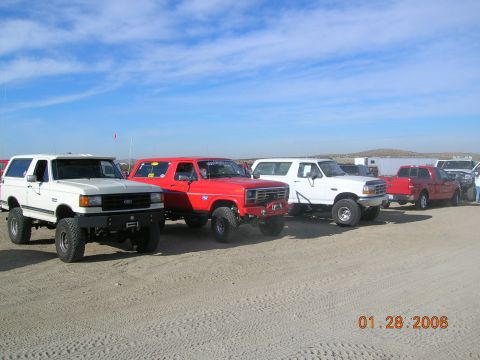 1985 Ford Bronco - A line of modified off-road vehicles, including Broncos and pickup trucks, are parked on a sandy terrain.