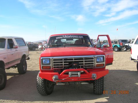 1985 Ford Bronco - A red Ford Bronco is parked in a sandy lot with other vehicles under a clear blue sky.