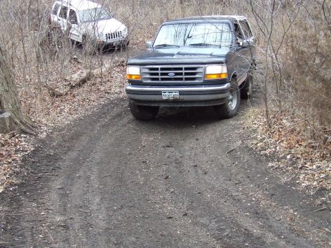 1994 Ford Bronco - Two SUVs navigate a wooded trail with fallen leaves on the ground.