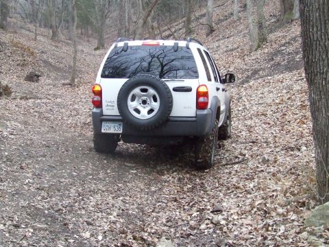 1994 Ford Bronco - A white Jeep Liberty drives off-road through a wooded area with fallen leaves on the ground.