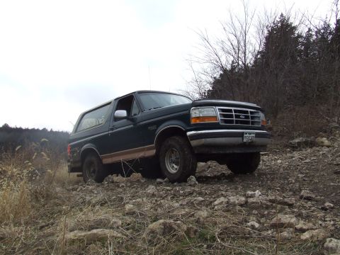 1994 Ford Bronco - A dark green 1994 Ford Bronco navigates a rough, rocky terrain with trees in the background.