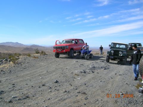 1985 Ford Bronco - Several off-road vehicles are parked on a dirt road in a desert landscape under a clear blue sky.