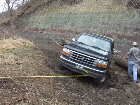 1994 Ford Bronco - A black 1994 Ford Bronco is stuck in the mud, with a person standing nearby and a yellow strap attached to the front.