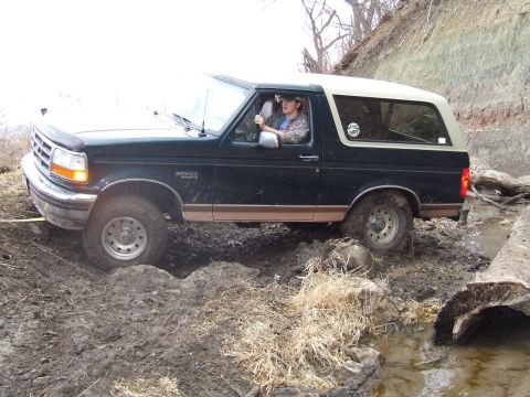1994 Ford Bronco - A Ford Bronco drives through a muddy, wooded area with a driver visible inside.