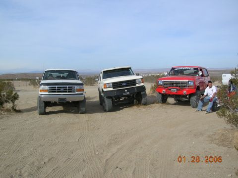 1985 Ford Bronco - Three modified Ford SUVs are parked in a desert landscape under a clear sky.