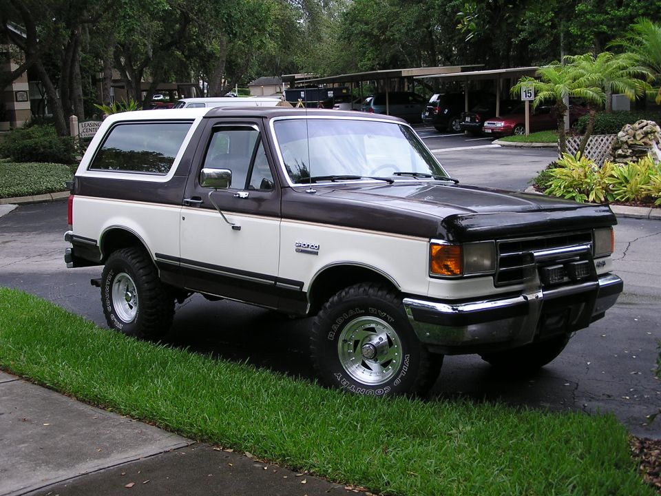 Two-tone brown and white Ford Bronco with white-letter tires parked on wet pavement near palm trees