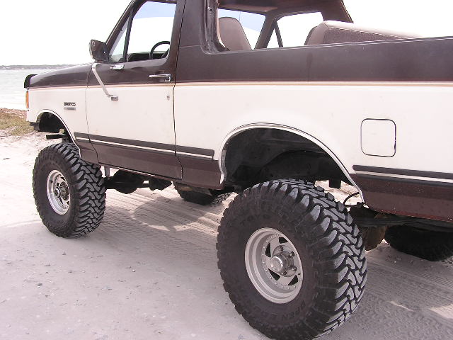 Side of lifted brown and tan Ford Bronco on tall mud tires parked on a beach