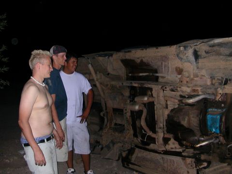 1984 Ford Bronco - Three men observe the heavily damaged and overturned chassis of a vehicle at night.