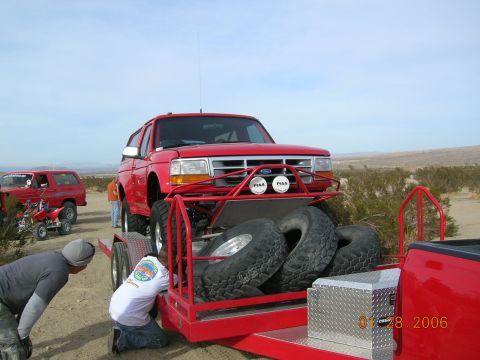 1985 Ford Bronco - A red Ford Bronco is loaded onto a trailer in a desert landscape, with other vehicles visible.