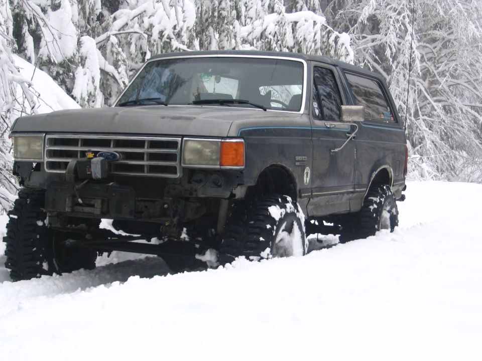 Gray 1987 Ford Bronco with large tires driving through deep snow in a wooded area