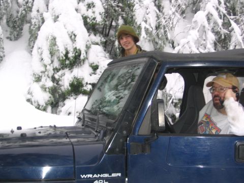 1987 Ford Bronco - Two people are inside a blue Jeep Wrangler, stopped in a snowy forest with snow-covered trees.