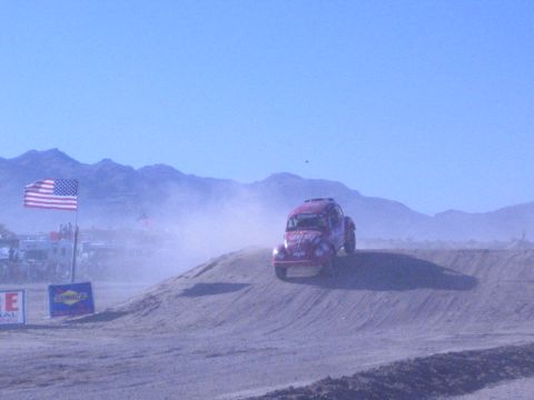 1990 Ford Bronco - red off-road racer crests a dirt berm in a dusty desert valley beside a US flag and a Sunoco sponsor banner.