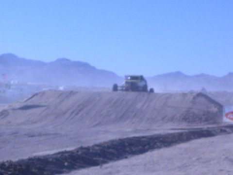 1990 Ford Bronco - yellow off-road buggy crests a distant dirt berm in a dusty, hazy desert valley.