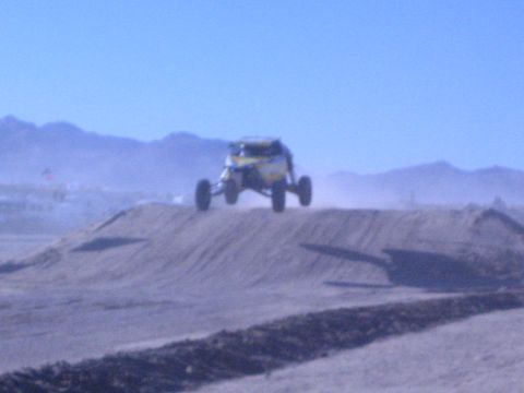 1990 Ford Bronco - yellow off-road buggy launches off a dirt jump, all four wheels in the air above a dusty desert course.