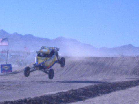 1990 Ford Bronco - blue-and-yellow off-road buggy catches air over a desert jump, kicking up dust beside a US flag.