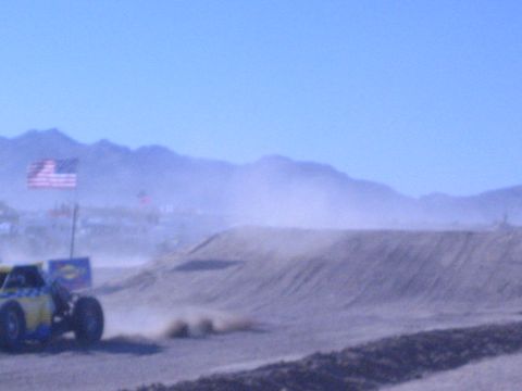 1990 Ford Bronco - blue-and-yellow off-road buggy churns up dust at the foot of a dirt berm near a US flag in the desert.