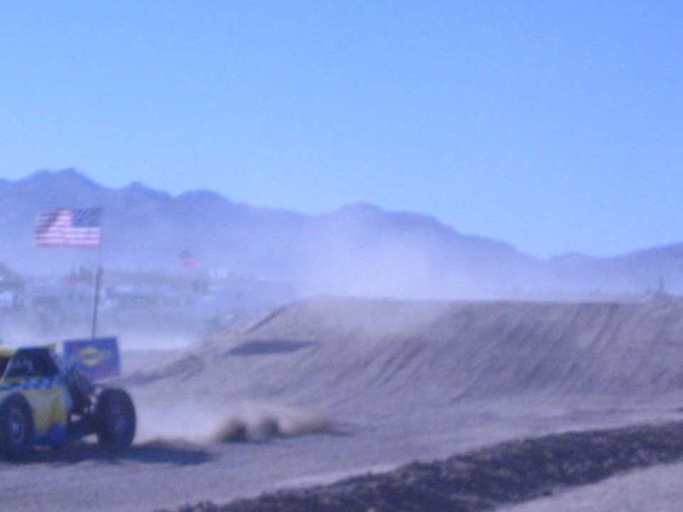 Blue and yellow off-road buggy throwing dust at the base of a dirt berm by a US flag
