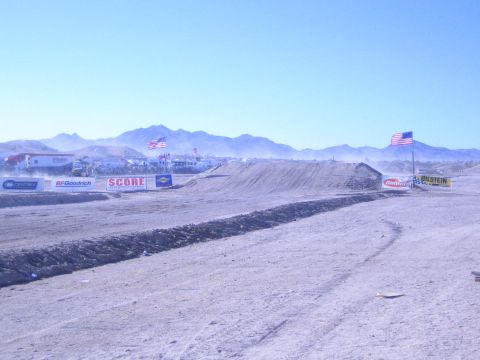 1990 Ford Bronco - wide desert off-road course lined with dirt berms, US flags and sponsor banners reading SCORE, BFGoodrich and Bilstein.