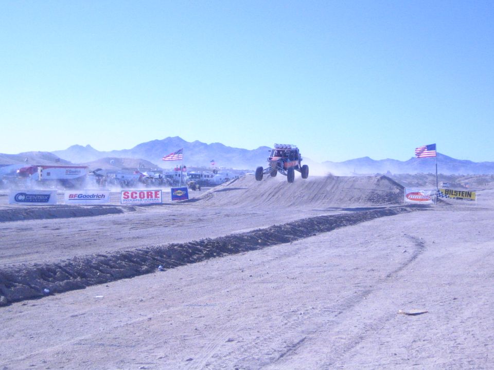 Off-road buggy airborne over a dirt jump on a desert course with US flags and SCORE banners