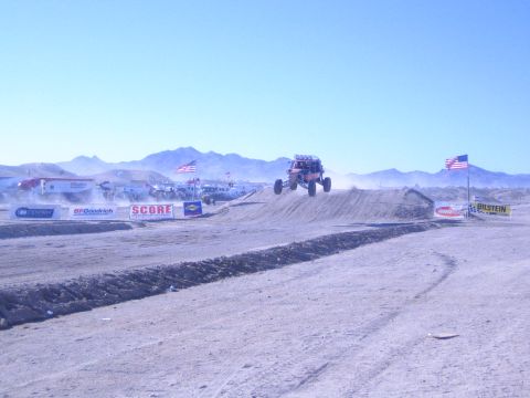 1990 Ford Bronco - off-road buggy catches air over a dirt jump on a desert course flanked by US flags and SCORE, BFGoodrich and Bilstein banners.