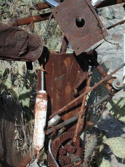 1994 Ford Bronco - Various heavily rusted mechanical parts are visible, including a shock absorber and gears, set against a backdrop of cacti and dry brush.