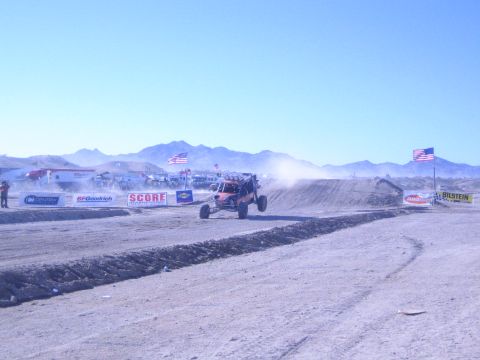 1990 Ford Bronco - open-wheel desert race buggy kicks up dust on a dirt course lined with sponsor banners and American flags under mountains.
