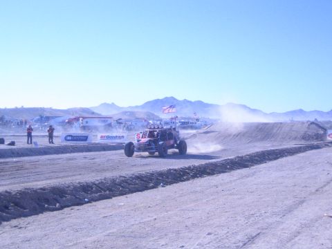 1990 Ford Bronco - single-seat off-road buggy races along a dirt track past BFGoodrich and Centric banners with US flags and desert mountains behind.