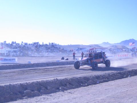 1990 Ford Bronco - orange-and-black desert buggy drives a dirt course as spectators line the hillside near a SCORE banner under a hazy sky.