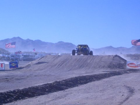 1990 Ford Bronco - off-road buggy catches air off a dirt jump on a desert race track flanked by large American flags and sponsor banners.