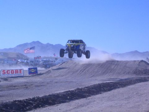 1990 Ford Bronco - yellow off-road buggy flies through the air off a dirt jump above a SCORE International banner with mountains behind.