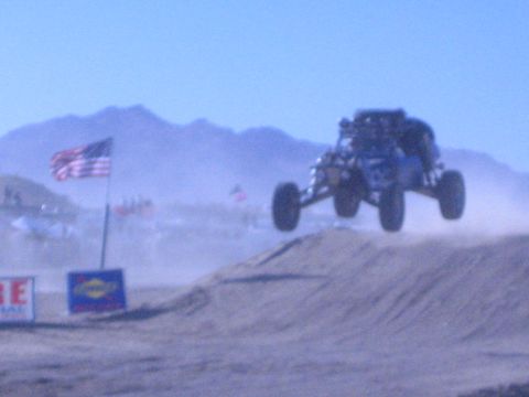1990 Ford Bronco - blue off-road buggy gets airborne over a dirt berm on a desert race course beside an American flag in a slightly blurred shot.