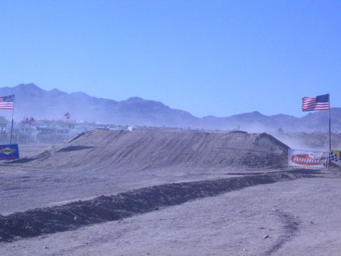 1990 Ford Bronco - graded dirt jump on a desert off-road course sits between American flags and sponsor banners under a clear blue sky.