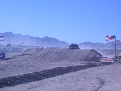 1990 Ford Bronco - small off-road vehicle crests a dirt jump in the distance on a dusty desert course lined with flags and banners.