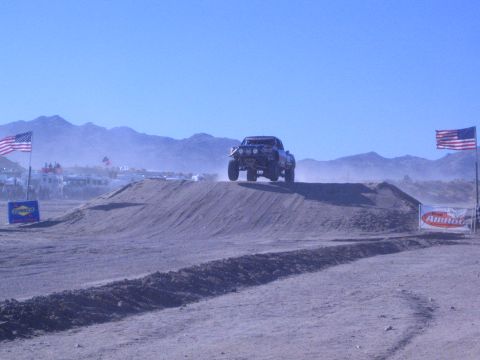 1990 Ford Bronco - off-road race truck launches over a dirt jump on a desert course between American flags and sponsor banners.