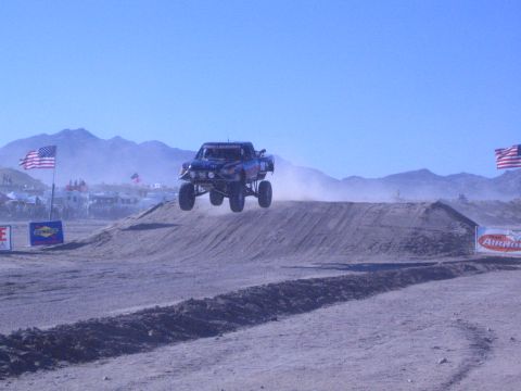 1990 Ford Bronco - blue off-road race truck gets air over a dirt berm on a desert track flanked by American flags and a Chevrolet-style banner.