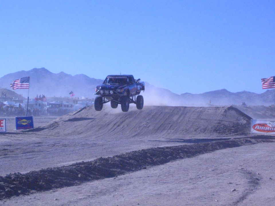 Blue off-road race truck jumping a dirt berm in the desert with flags