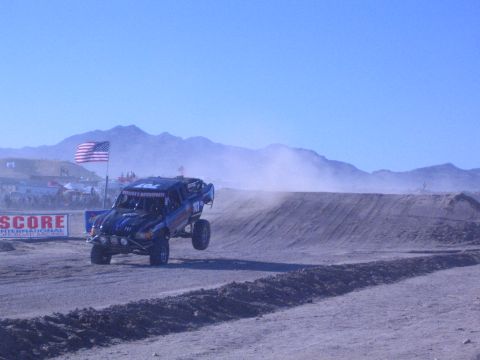 1990 Ford Bronco - blue off-road race truck drives a dirt desert course past a SCORE International banner and an American flag.