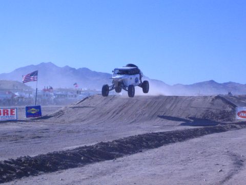 1990 Ford Bronco - white off-road buggy flies off a dirt jump on a desert race track lined with banners, flags and distant mountains.