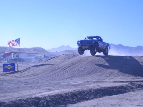 1990 Ford Bronco - blue off-road race truck gets airborne over a dirt berm on a desert track next to a Sunoco banner and US flag.