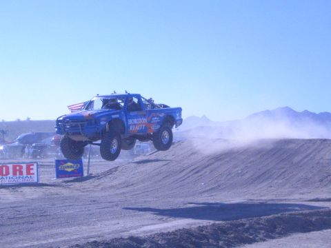 1990 Ford Bronco - blue off-road race truck lettered Horizon Realty jumps a dirt berm on a desert course near SCORE and Sunoco banners.