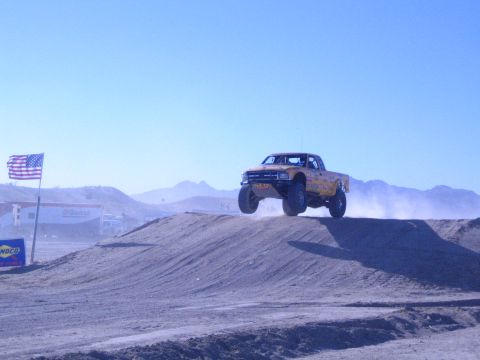 1990 Ford Bronco - yellow off-road race truck catches air off a dirt jump on a desert course with an American flag and mountains behind.