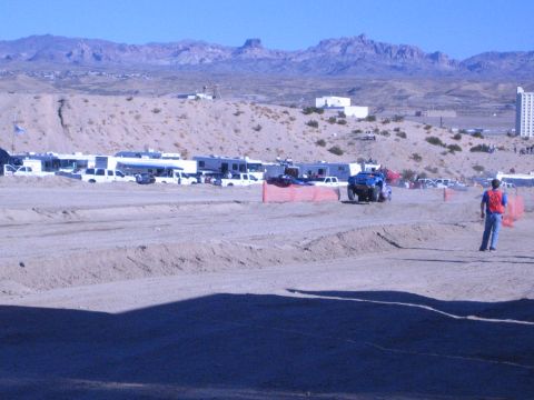 1990 Ford Bronco - blue race vehicle rounds a dirt track berm as RVs and pickups line the paddock and a flagger in red stands trackside.