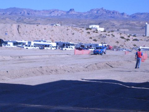 1990 Ford Bronco - blue off-road truck runs a dirt course past a row of RVs and pickups with a red-vested flagger watching from the berm.