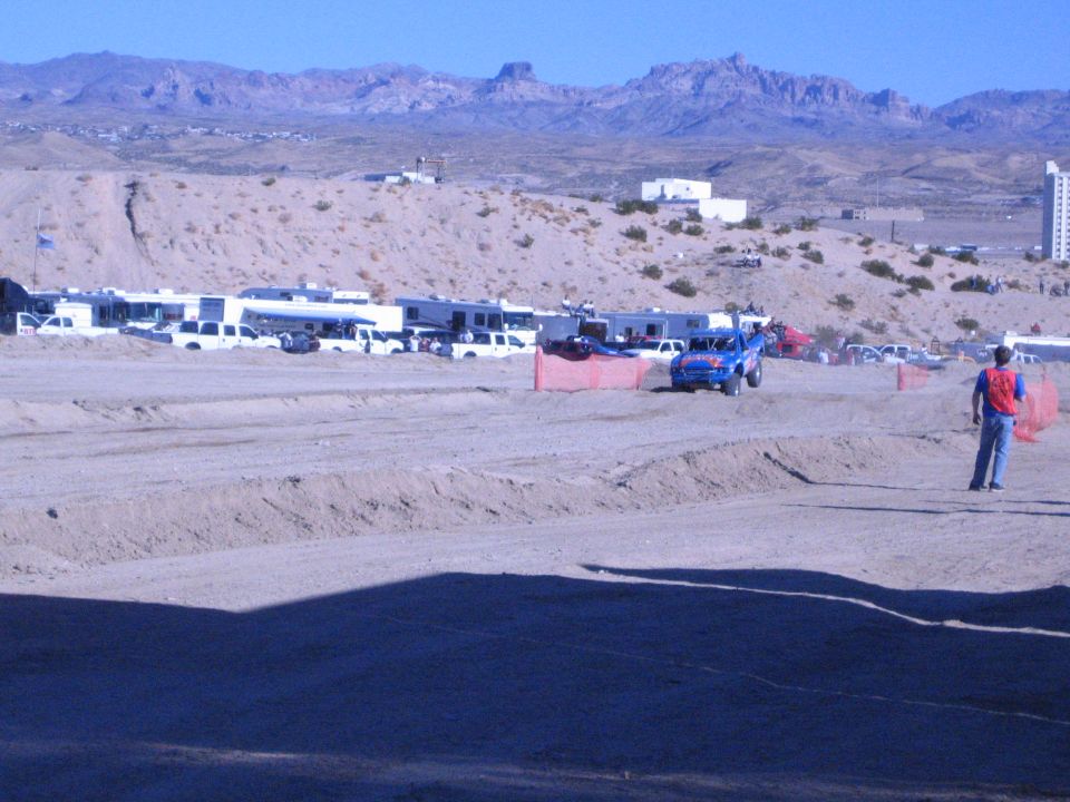 Blue off-road truck on a desert dirt track past RVs with a trackside flagger