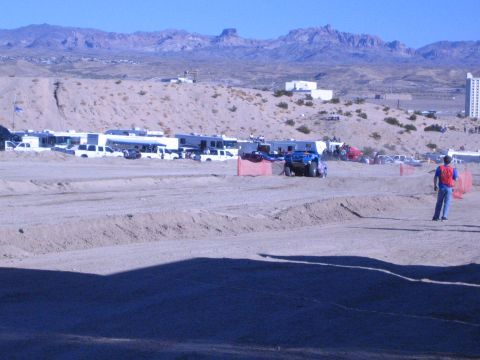 1990 Ford Bronco - blue off-road truck drives a graded dirt course in front of a paddock of RVs and trucks with a flagger standing trackside.