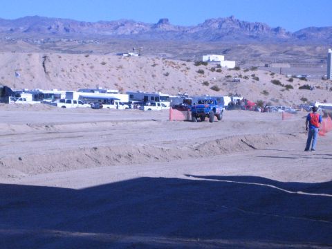 1990 Ford Bronco - blue off-road race truck moves along a dirt course in front of a long line of RVs and pickups under desert mountains.